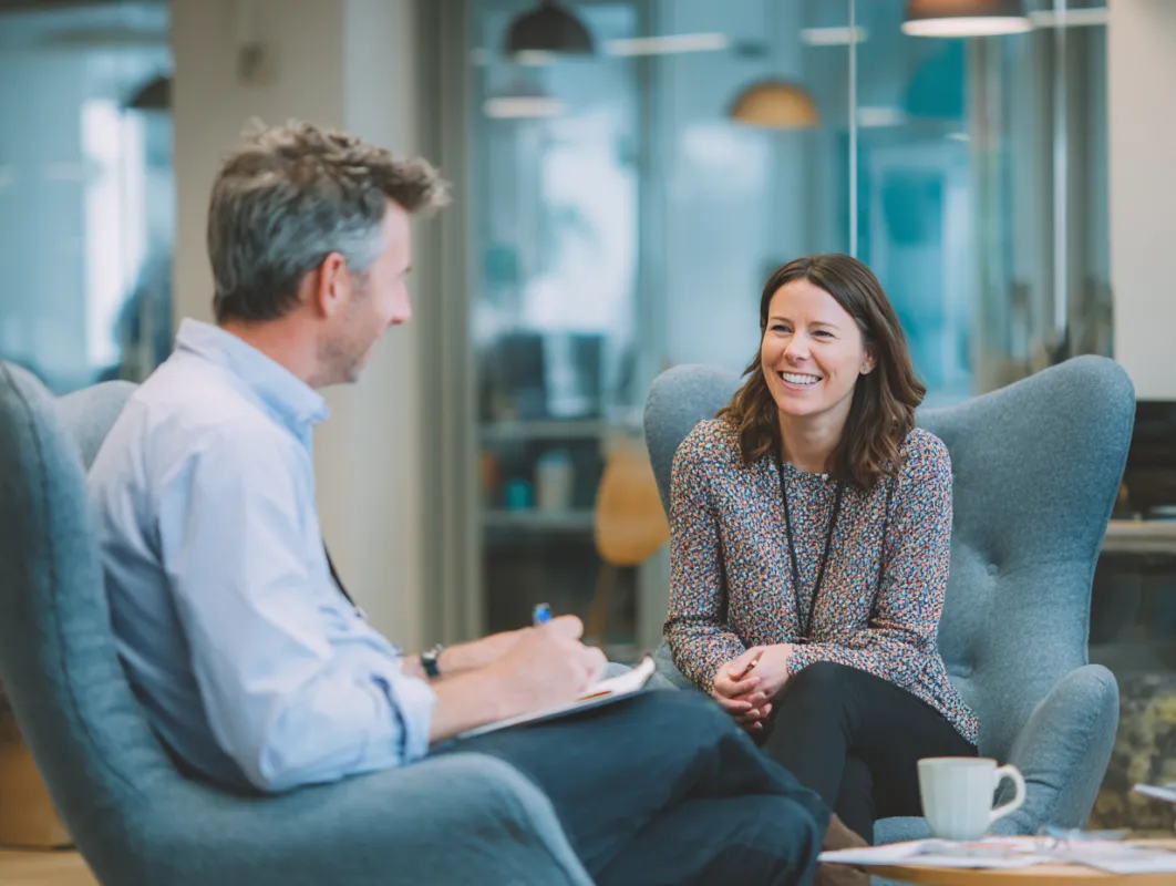 Two professionals having a conversation in a modern office setting with glass walls and pendant lighting.