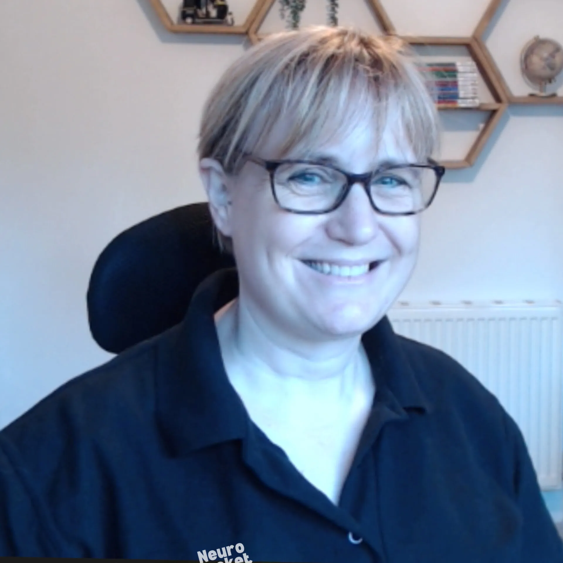 Picture of Kerry with short light hair and glasses smiling at camera in home office with hexagonal shelves behind her.