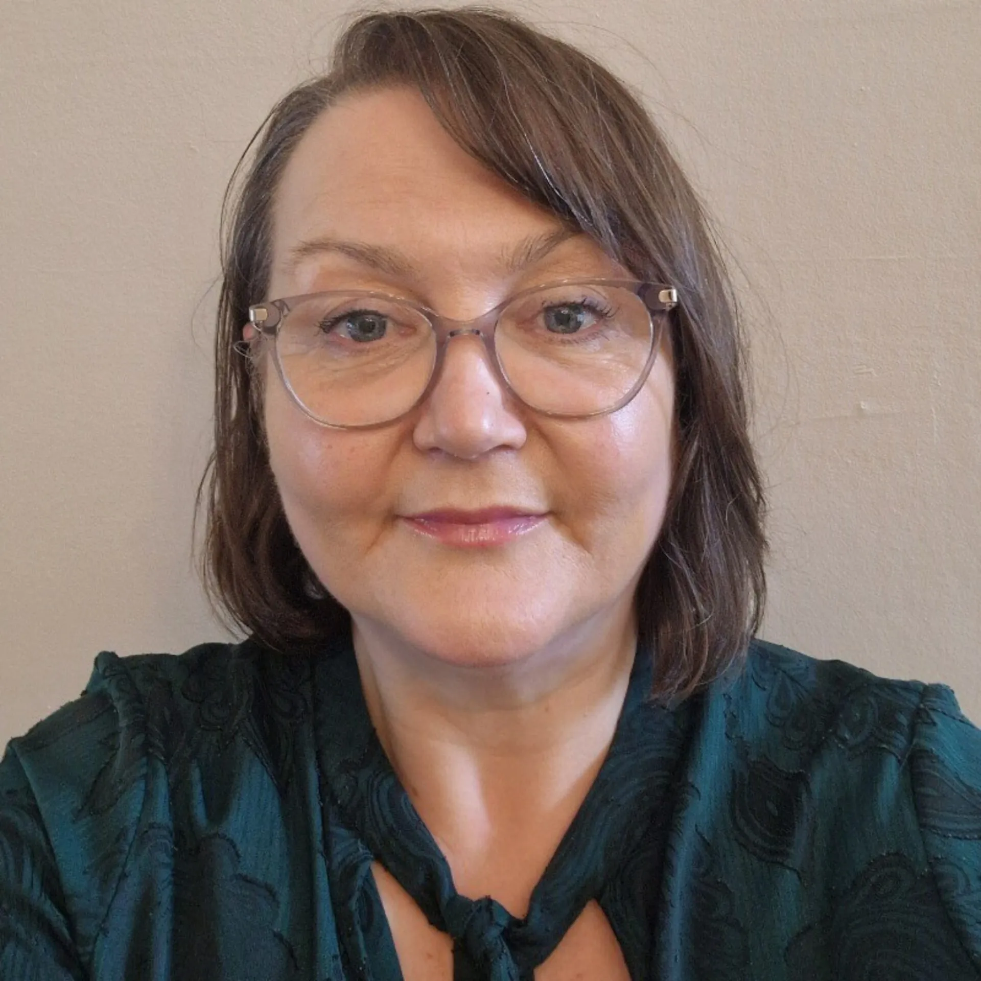 Woman with shoulder-length brown hair wearing clear-framed glasses and a dark teal patterned shirt against a neutral beige background.