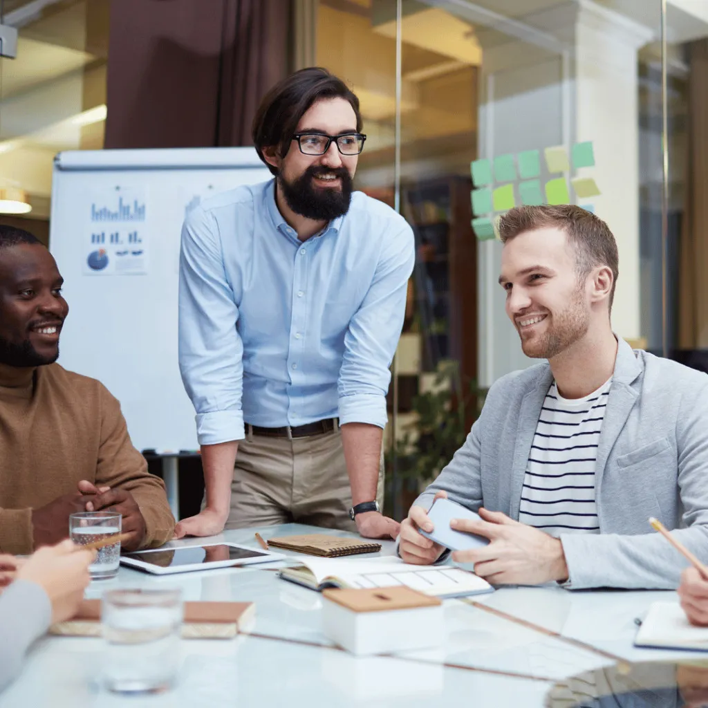 Three diverse professionals collaborating at a table during a business meeting in a modern office with whiteboards and sticky notes visible.